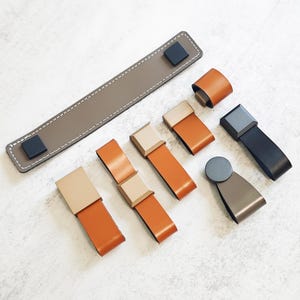 Könnte beinhalten: Eine Auswahl an Leder-Schrankgriffen und -Ziehgriffen in verschiedenen Stilen und Farben. Die Auswahl umfasst einen langen, rechteckigen Griff in Taupe mit schwarzen Quadraten und mehrere kürzere Griffe in Orange, Beige und Schwarz mit metallischen Akzenten. Das Möbelzubehör.