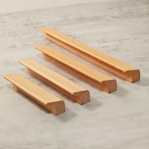 Könnte beinhalten: Vier Holzschrankgriffe in verschiedenen Längen, diagonal angeordnet. Die Griffe haben eine helle, natürliche Holzfarbe mit rechteckigem Profil und einem kleinen, erhöhten Griff. Die Holzmaserung ist sichtbar, vor neutralem Hintergrund.