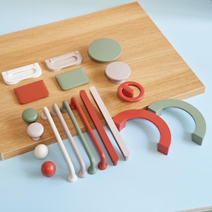 Könnte beinhalten: Eine Sammlung farbenfroher Schrankbeschläge, darunter Knöpfe, Griffe und Ziehgriffe in verschiedenen Formen und Größen. Die Beschläge sind in Weiß-, Rot-, Grün- und Beigetönen auf einer Holzoberfläche angeordnet.