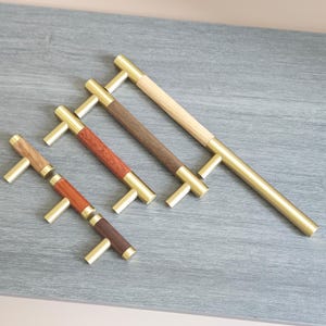 Può includere: Quattro maniglie per mobili con ferramenta in ottone e manici in legno in varie tonalità di marrone. Le maniglie presentano diverse venature e finiture del legno, tra cui toni chiari, medi e scuri. Le maniglie sono disposte diagonalmente su una superficie grigia.