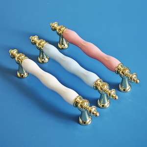 Op de afbeelding: Vier kastgrepen met een gouden afwerking en kleurrijke keramische centra. De handgrepen zijn wit, lichtblauw en roze. De handgrepen liggen op een blauw oppervlak.