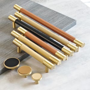 Op de afbeelding: Een collectie kastbeslag met messing handgrepen en knoppen. De handgrepen hebben een cilindrisch ontwerp met leren of houten middenstukken in verschillende kleuren, waaronder bruin, zwart en beige. De knoppen zijn rond met bijpassende afwerkingen.