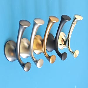May include: Five metal wall hooks in silver, gold, black, and brass finishes. Each hook has a curved design with a rounded top and a circular base. The hooks are mounted on a blue wall. The design is suitable for a hallway.