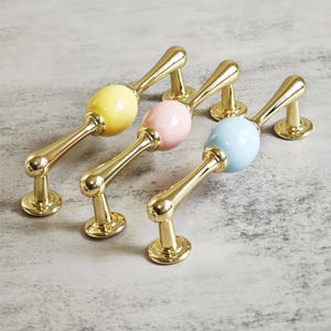 Op de afbeelding: Vier kastgrepen met een gouden afwerking en een kleurrijk keramisch middenstuk. De grepen zijn geel, roze en blauw. De grepen hebben een gebogen ontwerp en staan tegen een lichtgrijze achtergrond.