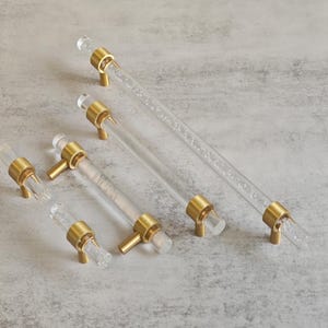May include: Clear acrylic cabinet handles with brass-coloured fittings. The handles are cylindrical with a bubble-like texture inside the acrylic. Various sizes are displayed, showcasing the design.