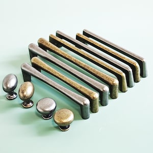 Op de afbeelding: Een verzameling kastbeslag, waaronder handgrepen en knoppen met een zilveren en antiek messing afwerking. De handgrepen zijn rechthoekig en de knoppen zijn ovaal en rond. De hardware is geplaatst op een lichtgroen oppervlak.