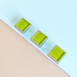 Puede incluir: Tres tiradores cuadrados para armarios con un centro de cristal verde brillante y un marco de metal plateado. Los tiradores están dispuestos en diagonal sobre un fondo azul claro y beige. Ideales para añadir un toque de color.