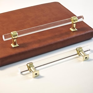 Könnte beinhalten: Zwei transparente Acryl-Schrankgriffe mit goldfarbenen Metallakzenten. Die Griffe haben einen zylindrischen Griff und liegen auf einer braunen Oberfläche. Die Hardware ist quadratisch mit einem zylindrischen Sockel.