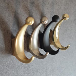 May include: Four decorative wall hooks in gold, silver, black, and brass. Each hook has a curved design with a rounded top and a curved hook. The hooks are mounted on a grey wall, offering a stylish storage solution.
