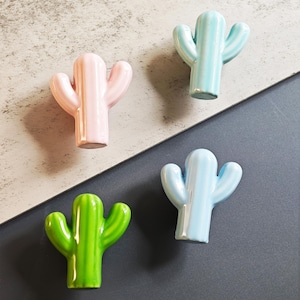 Può includere: Quattro statuette di cactus in ceramica dai colori pastello: rosa, azzurro, verde e blu. Ogni cactus ha una forma unica e una finitura lucida. Le statuette sono disposte su uno sfondo diagonale grigio e bianco.