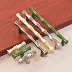 May include: A selection of bamboo-style cabinet pulls in various colours, including green, silver, gold, and bronze. The pulls are arranged on a wooden surface, showcasing their design and finish. These decorative hardware pieces add a natural touch to furniture.