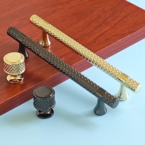 Op de afbeelding: Twee kastgrepen en twee knoppen in goud en zwart. De grepen hebben een diamantpatroon en zijn gemonteerd op een gouden of zwarte basis. De knoppen zijn rond met een diamantpatroon en zijn goud of zwart.