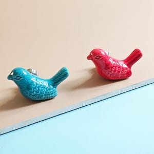 May include: Two ceramic bird-shaped cabinet knobs. One is turquoise, the other is red. Both have a glossy finish and are mounted on a silver-colored metal base. The knobs are designed to add a decorative touch to furniture.