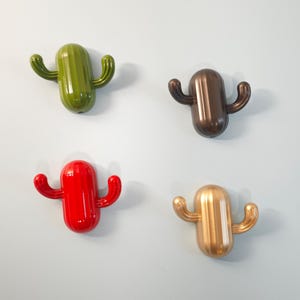 Può includere: Quattro ganci da parete a forma di cactus in verde, bronzo, rosso e oro. Ogni gancio ha un corpo arrotondato con due braccia curve, che assomigliano a un cactus. I ganci sono lucidi e montati su una parete azzurra.
