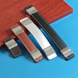 May include: Four leather cabinet pulls in different sizes and colours: dark brown, light brown, white, and black. Each pull has a silver-toned metal accent and white stitching. The pulls are arranged on a wooden surface.