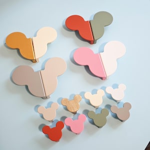 Op de afbeelding: Een verzameling houten kastknoppen in verschillende kleuren, waaronder oranje, rood, roze, grijs en wit. De knoppen zijn in de vorm van een muizenkop en zijn gerangschikt op een lichtblauwe ondergrond.