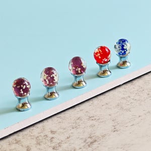 May include: Five decorative cabinet knobs with a spherical design. Each knob has a clear, round top filled with glitter and set on a silver-coloured base. The tops are in shades of red, purple, and blue, offering a colourful accent.