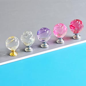 May include: Five decorative cabinet knobs with a rose design. The knobs are made of clear glass in various colors, including clear, purple, pink, and red. The bases are silver, except for one gold base. These knobs are a decorative hardware item.