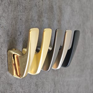 Può includere: Una fila di cinque ganci in metallo in varie tonalità di oro, argento e nero. I ganci sono montati su una superficie grigia. I ganci hanno un design curvo e moderno.