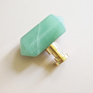 Puede incluir: Un tirador de armario de piedra preciosa verde claro y facetada con una base de metal dorado. La piedra preciosa es rectangular con bordes angulados. El tirador está diseñado para muebles y decoración del hogar.