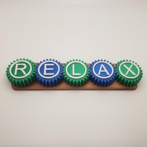 May include: Five colorful gears spelling out the word "RELAX" in white letters. The gears are green, blue, and green, with a brown wooden base.