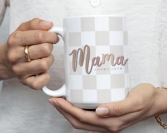 May include: White ceramic mug with a checkered pattern in beige and white. The word "Mama" is written in a cursive font in a rose gold color. The text "LOOMA 18TH" is below the word "Mama".