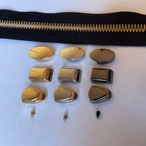 May include: Assortment of zipper pulls in gold, silver, and black metal finishes. The pulls are in various shapes, including oval, rectangular, and teardrop. Two small screws are also included.