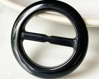 May include: A black plastic buckle with a smooth, shiny finish. The buckle has a round shape with a center opening and a horizontal bar across the middle.