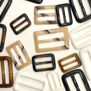 May include: Assortment of plastic buckle closures in various colours and patterns, including brown, black, white, and beige. The buckles are rectangular with two parallel slots.