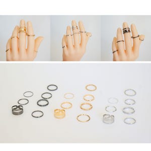May include: Set of 18 gold, silver, and black rings. The rings are various styles and sizes, including thin bands, thicker bands, and rings with textured details.