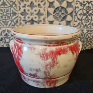 Peut inclure: Un pot en céramique blanche avec un glaçage rouge, présentant un aspect vieilli. Le pot a deux poignées sur les côtés et une forme arrondie.