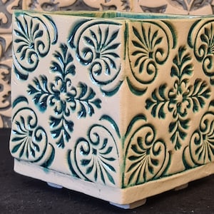 May include: A square, off-white ceramic planter with a raised, teal floral pattern. The planter has a rectangular shape and sits on small, square feet. The floral design is repeated across all sides of the planter.