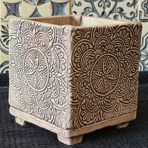 Könnte beinhalten: Quadratischer, beigefarbener Blumentopf mit aufwendigen, geprägten Blumenmustern. Der Topf hat ein leicht gealtertes Aussehen und steht auf vier kleinen Füßen. Das Design zeigt symmetrische, mandalaartige Motive und erzeugt eine dekorative und strukturierte Oberfläche.