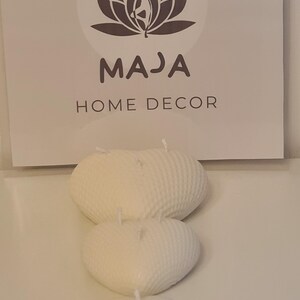 Peut inclure: Trois bougies blanches en forme de cœur avec une surface texturée. Les bougies sont disposées dans un ordre de taille décroissante, la plus grande bougie en haut et la plus petite bougie en bas. Les bougies sont sur une surface blanche. L'image comprend également un logo pour "MAJA HOME DECOR" avec un motif de fleur de lotus.