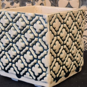 Op de afbeelding: Een vierkante keramische plantenpot met een witte glazuur en een blauw geometrisch patroon. De pot heeft een licht verhoogde rand en vier kleine voetjes aan de onderkant.