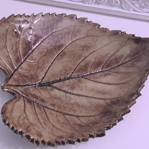 Pode incluir: Um prato de cerâmica em forma de folha com um esmalte castanho e veios detalhados. O prato tem uma borda festonada e é perfeito para servir petiscos ou pequenos itens.