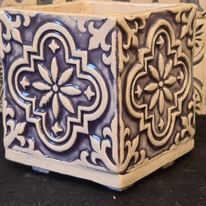 Könnte beinhalten: Ein quadratischer Keramik-Pflanztopf mit weißer Glasur und einem violetten Design. Der Topf hat ein geometrisches Muster mit einem floralen Motiv.