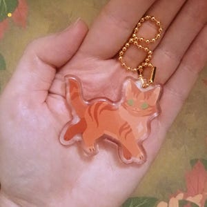 Pu&ograve; includere: Un ciondolo a forma di gatto in acrilico trasparente con un design tabby arancione e occhi verdi. Il ciondolo &egrave; attaccato a una catena a sfera e a una chiusura color oro. Il gatto cammina con la coda alzata. Lo sfondo &egrave; un motivo floreale.