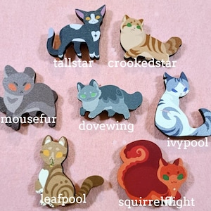 May include: A collection of seven wooden cat-shaped pins in various colors and patterns. Each pin features a different cat design with names like "tallstar" and "crookedstar" printed below. The pins are arranged on a pink surface.
