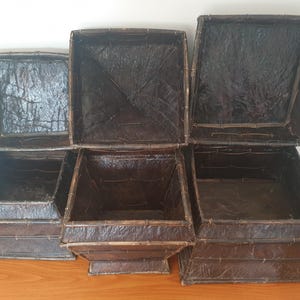 May include: A set of three square dark brown containers with lids, constructed from a textured material with a visible bamboo-like frame. Each container features a tapered base and a matching lid, displaying a deep, glossy finish throughout.