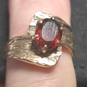 Könnte beinhalten: Goldfarbener Ring mit einem großen, oval geschliffenen, tiefroten Edelstein. Der Ring hat ein strukturiertes, rindenartiges Design auf dem Band und der Fassung, was eine rustikale und organische Ästhetik erzeugt. Der Edelstein wird von Zinken gehalten.