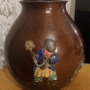 May include: A brown ceramic vase with a textured finish. The vase is decorated with a relief of a figure in a blue coat, yellow skirt, and red scarf holding a fan. The vase has a rounded body and a slender neck.