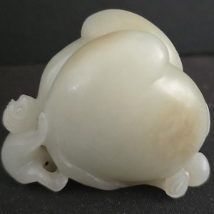 May include: A carved, off-white sculpture of a fruit, possibly a peach, with a small figure clinging to its side. The sculpture has a smooth, polished surface with subtle variations in color.