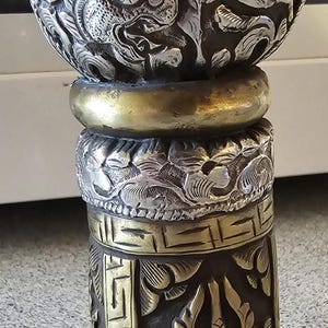 May include: A cylindrical metal object featuring a rounded top with intricate relief carvings of a creature. The body displays a geometric Greek key border, a central crescent moon and flame motif, and a textured silver and brass finish throughout.