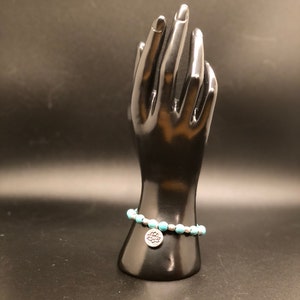 Può includere: Un braccialetto di perline turchesi con un ciondolo d'argento a forma di fiore di loto.