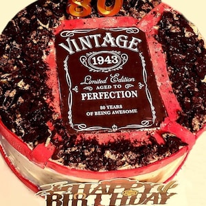 May include: A red and white cake decorated with a black and white label that says "Vintage 1943 Limited Edition Aged to Perfection 80 Years of Being Awesome". The cake is also decorated with chocolate sprinkles and a gold number 80 candle.