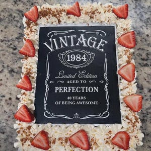 May include: A square cake decorated with white frosting and strawberries. The cake has a black fondant topper with white text that reads "Vintage 1984 Limited Edition Aged to Perfection 40 Years of Being Awesome".