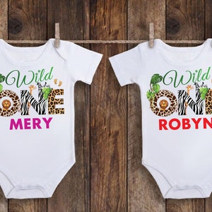May include: Two white baby onesies with a jungle theme. Each onesie has the words "Wild One" in green script, with the number "ONE" in a zebra print, leopard print, and a brown circle with a lion. One says "MERY" in pink, the other says "ROBYN" in red.