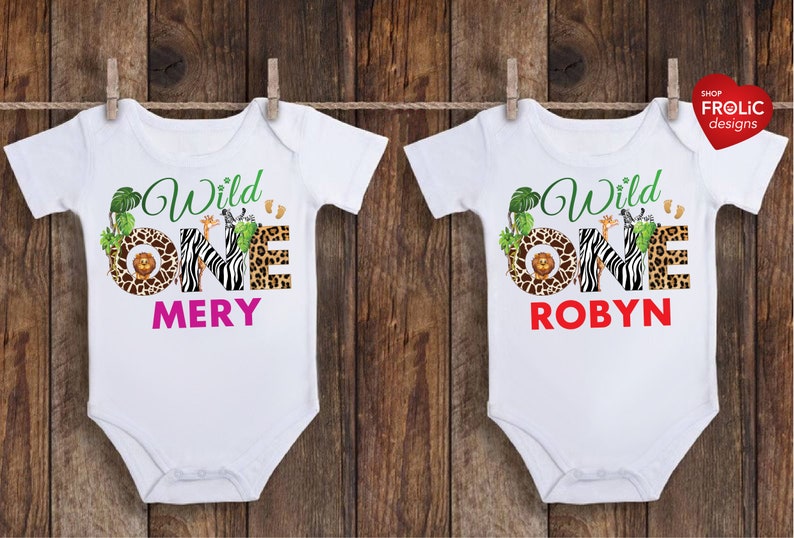 May include: Two white baby onesies with a jungle theme. Each onesie has the words "Wild One" in green script, with the number "ONE" in a zebra print, leopard print, and a brown circle with a lion. One says "MERY" in pink, the other says "ROBYN" in red.