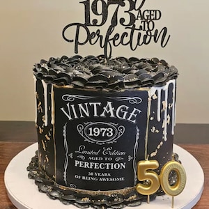 May include: Black and gold cake with a vintage-style label that reads "Vintage 1973 Limited Edition Aged to Perfection 50 Years of Being Awesome". The cake is decorated with black frosting and gold accents. A black and gold topper reads "Since 1973 Aged to Perfection". A gold candle shaped like the number 50 is on top of the cake.